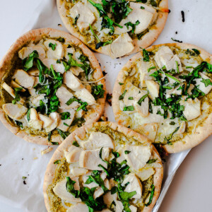 Four chicken pesto pita pizzas baking on a sheet pan, topped with pesto, mozzarella, sliced chicken, and chopped greens.