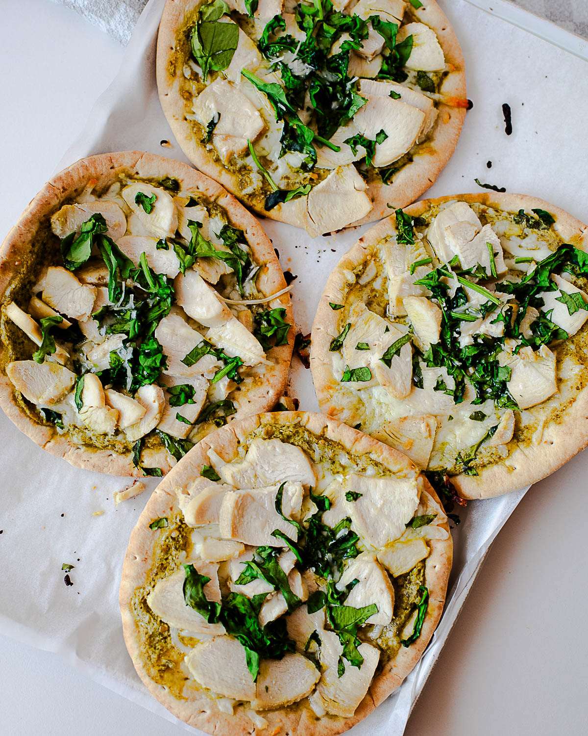 Four chicken pesto pita pizzas baking on a sheet pan, topped with pesto, mozzarella, sliced chicken, and chopped greens.