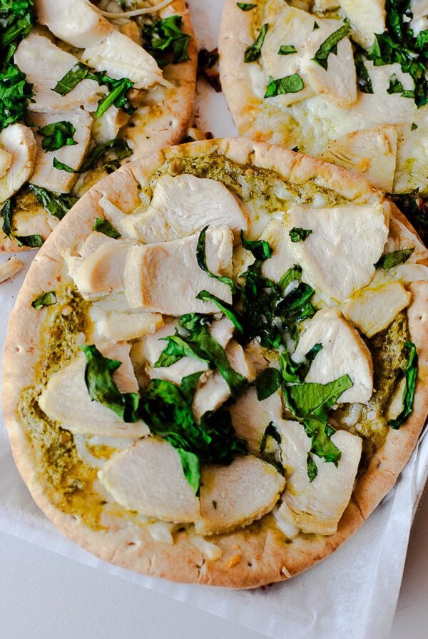 Close-up of a chicken pesto flatbread pizza showing melted mozzarella, pesto sauce, sliced chicken, and fresh basil on a crisp pita crust.