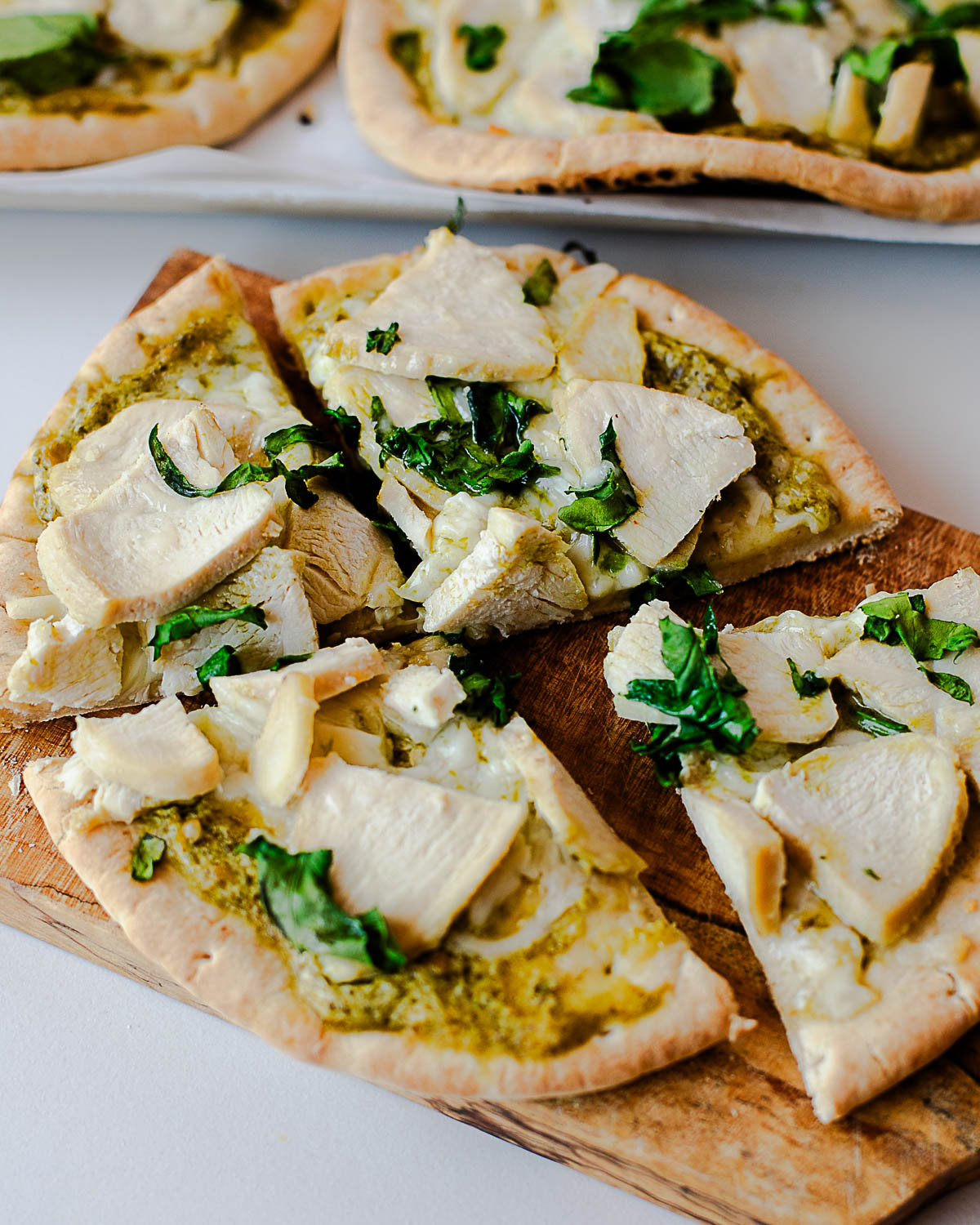 Chicken pesto pita pizza sliced into quarters on a wooden cutting board, with pesto sauce swirled under melted mozzarella and tender chicken pieces on top.