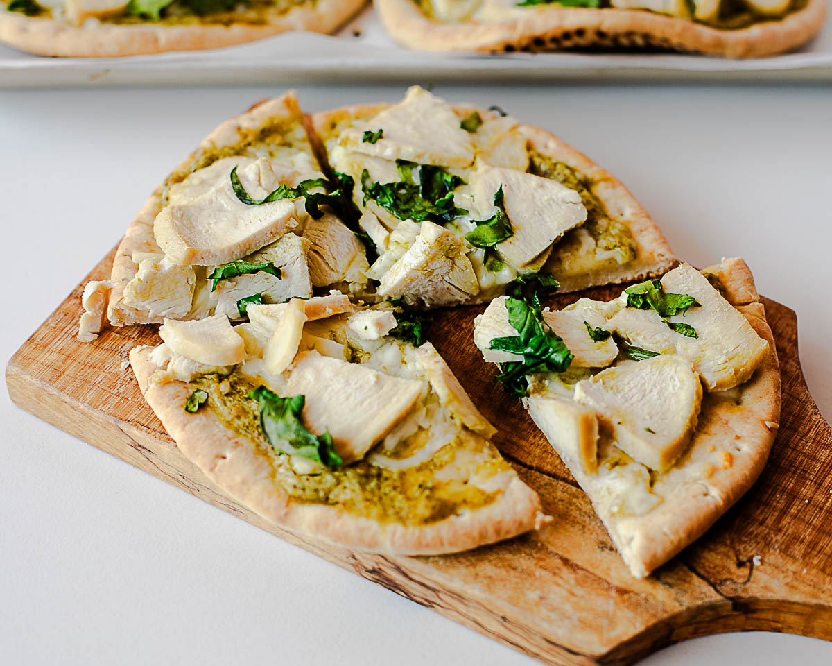 Chicken pesto flatbread pizza sliced into wedges on a wooden board with extra pizzas in the background.