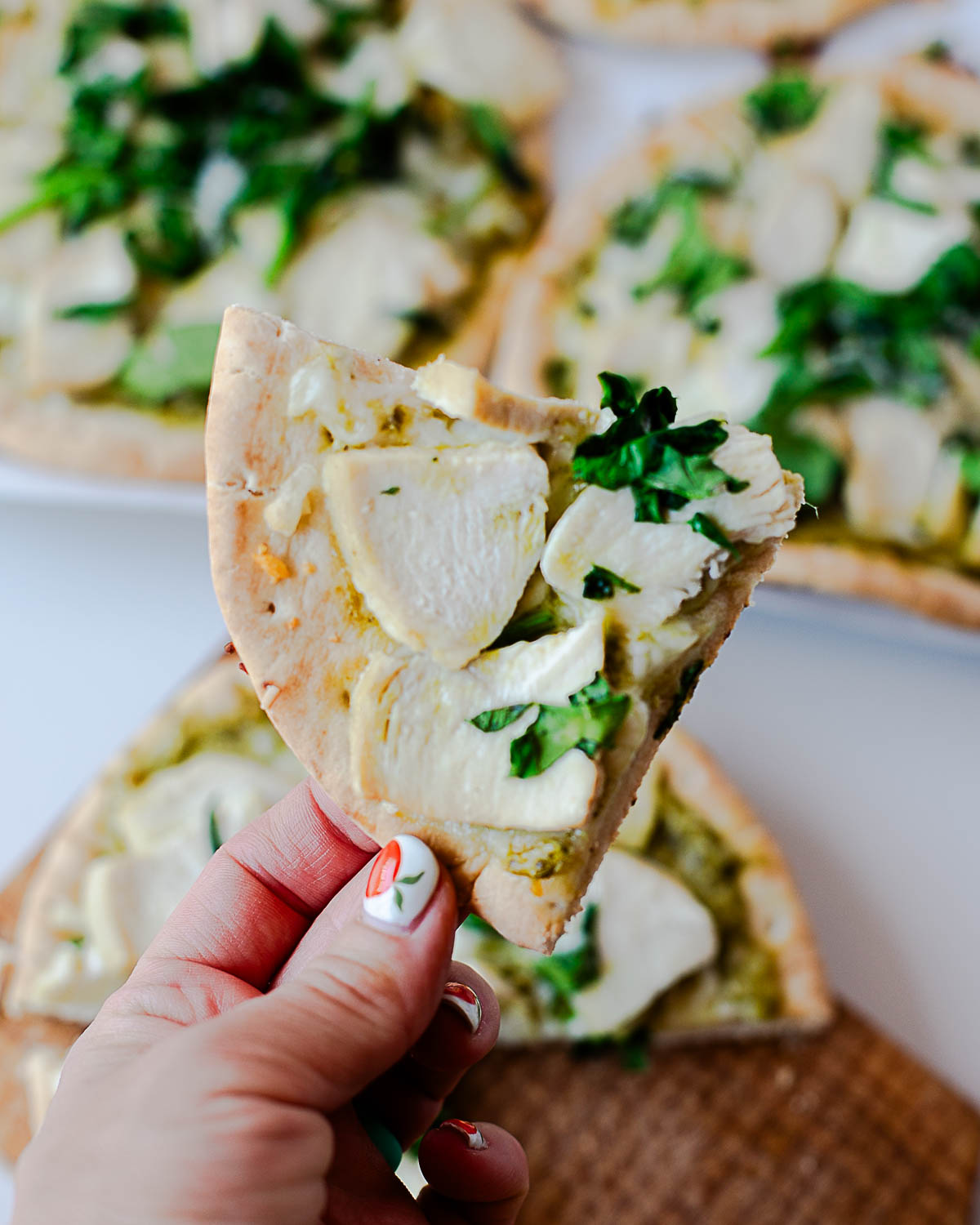 Hand holding a slice of chicken pesto flatbread pizza with melted mozzarella and pesto, with the rest of the pizzas blurred in the background.