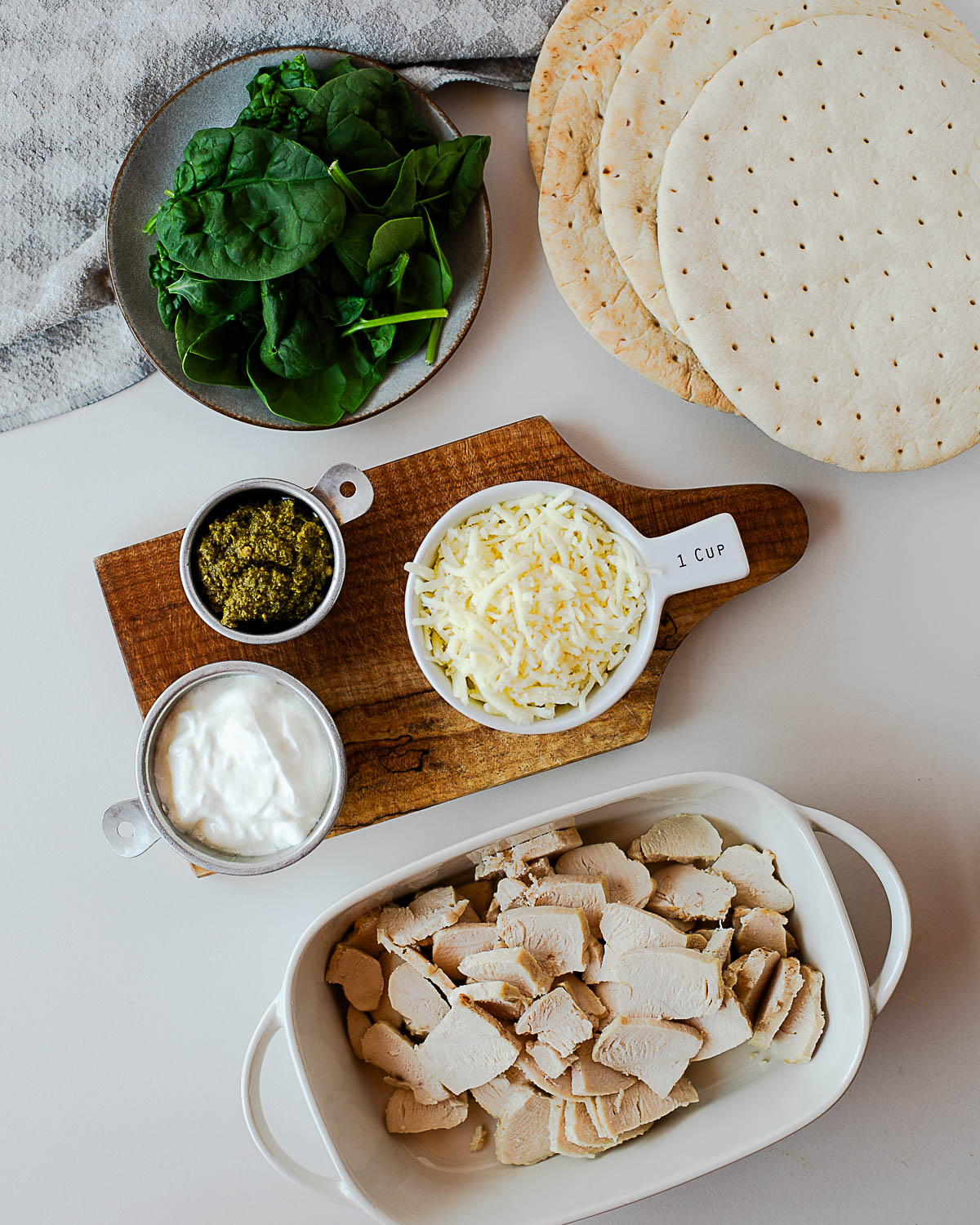 Ingredients for chicken pesto flatbread pizzas including pita rounds, cooked chicken, fresh spinach, pesto, Greek yogurt, and shredded mozzarella on a countertop.