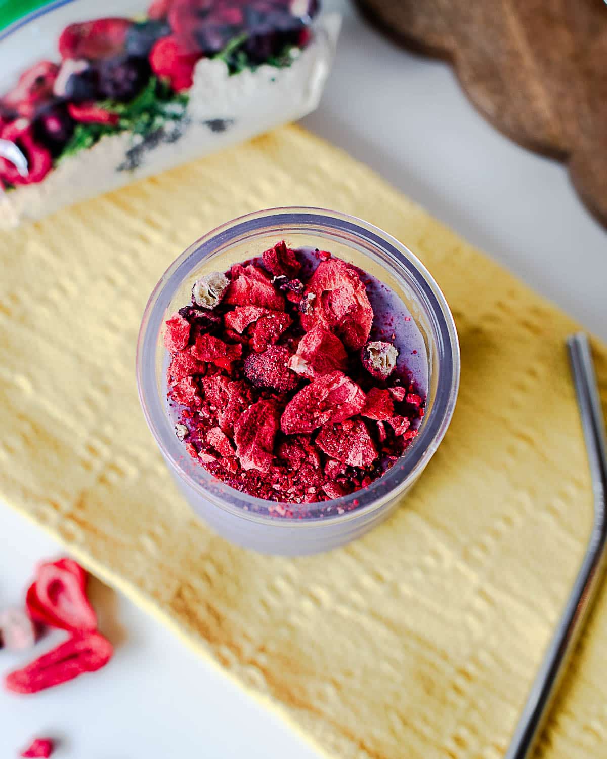 Top-down view of a purple berry protein smoothie topped with crushed freeze-dried berries on a yellow towel, with a smoothie pack in the background.