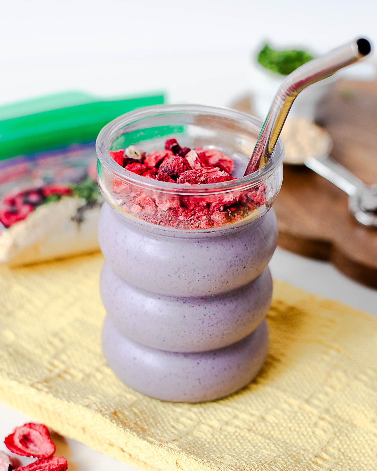 Purple berry protein smoothie in a ridged glass with a metal straw and crushed freeze-dried berry topping, with smoothie pack ingredients blurred behind.