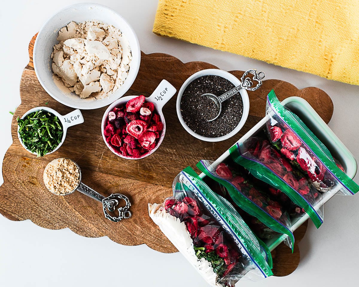 Measured ingredients for pantry smoothie packs—protein powder, chia seeds, peanut butter powder, freeze-dried berries, and spinach flakes—next to several filled zip-top bags in a dish.