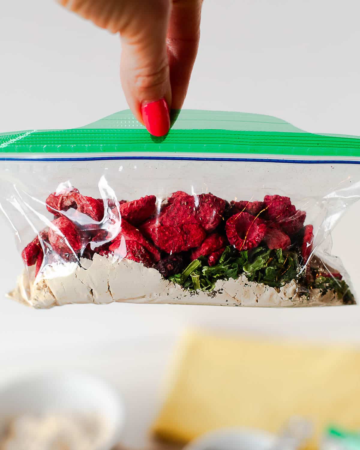 Hand holding a finished pantry smoothie pack in a zip-top bag, showing layers of protein powder, freeze-dried berries, and spinach flakes.