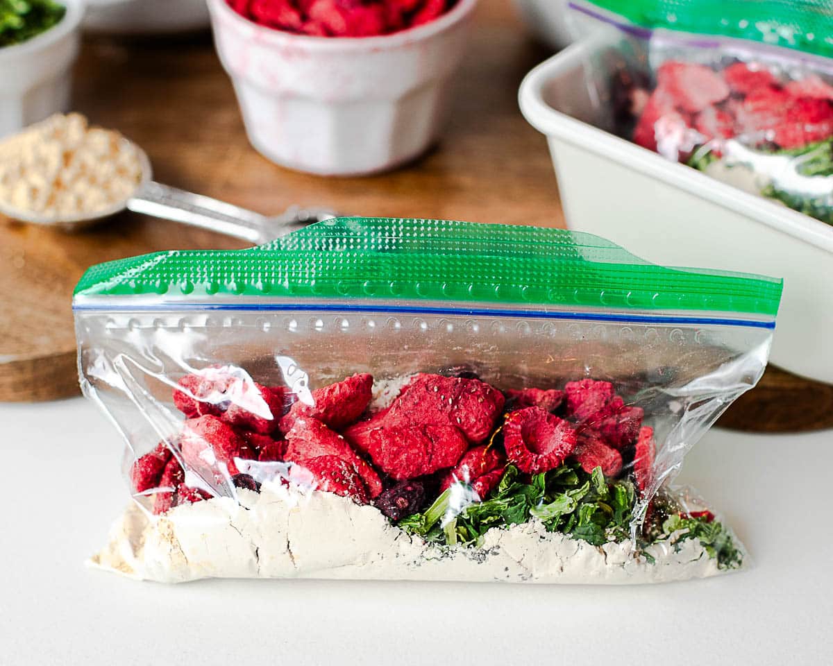 Single pantry smoothie pack in a zip-top bag filled with protein powder, freeze-dried berries, and spinach flakes on a white surface.