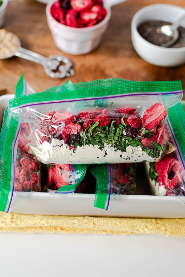 Close-up of a zip-top smoothie pack filled with protein powder, freeze-dried berries, and spinach flakes with more ingredients in the background.