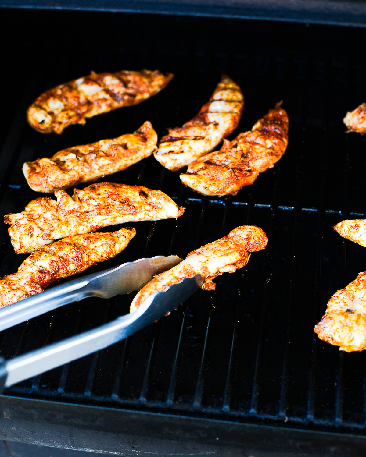 Close-up of Traeger chicken tenders on the grill as they cook over open grates.
Tongs flipping seasoned chicken tenders on the Traeger grill.
