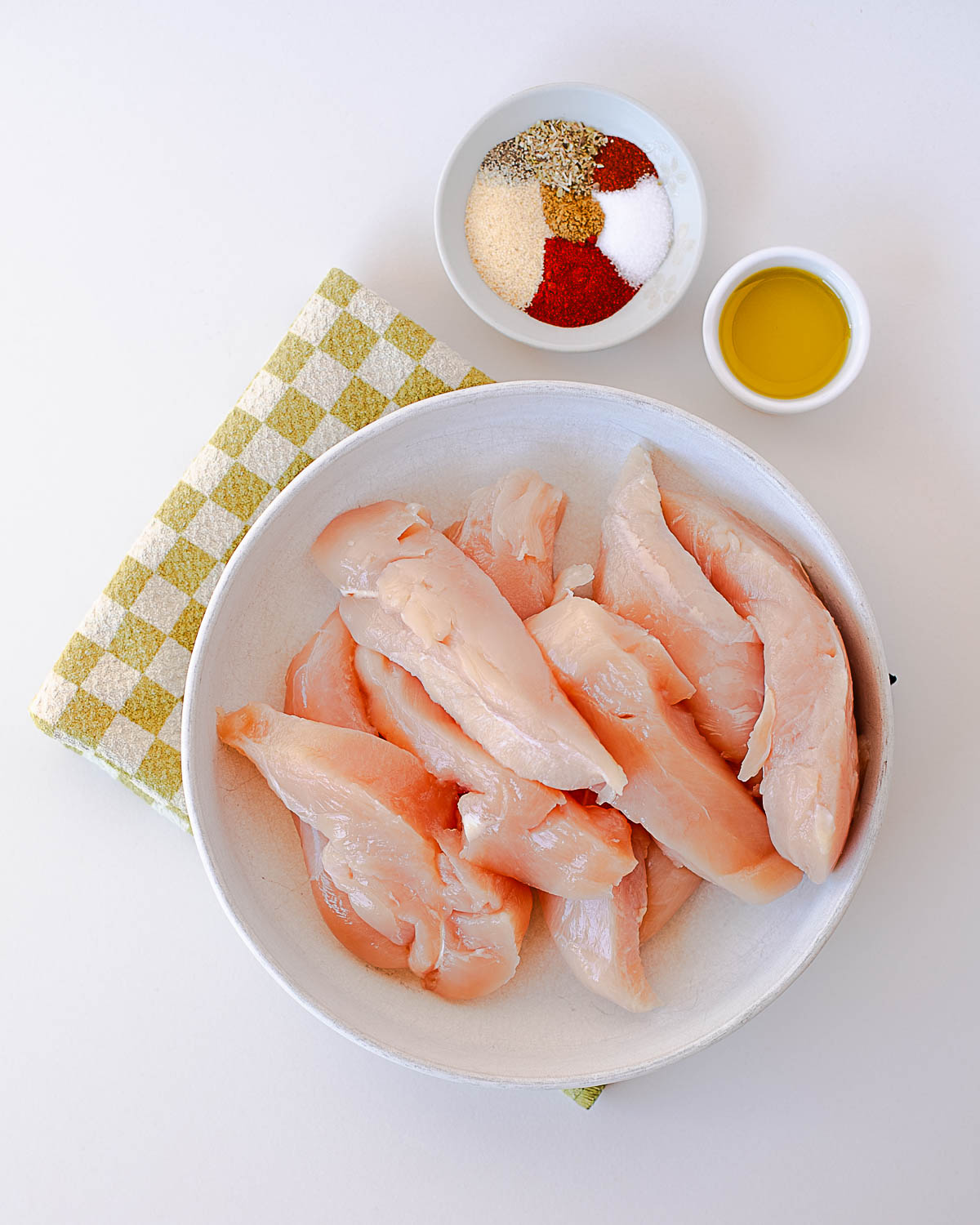 Raw chicken tenderloins with a small bowl of spices and olive oil beside them for seasoning.