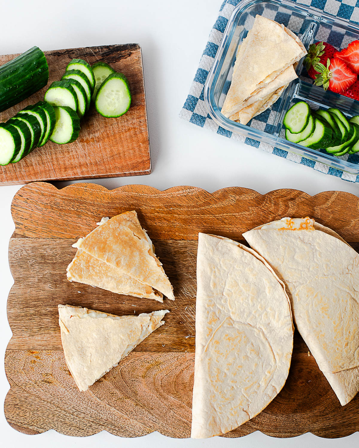 Tuna melt quesadilla wedges served on a cutting board with sliced cucumbers and strawberries in the background.