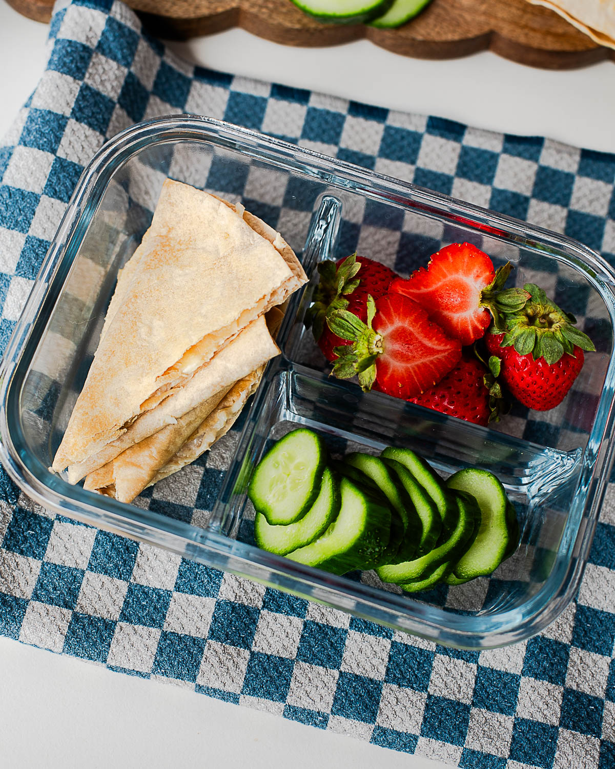 Lunchbox-style tuna melt quesadilla meal with sliced cucumbers and strawberries on a blue checkered towel.