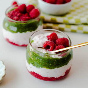 Styled raspberry matcha overnight oats parfaits arranged on a white background with fresh raspberries and kitchen linens.