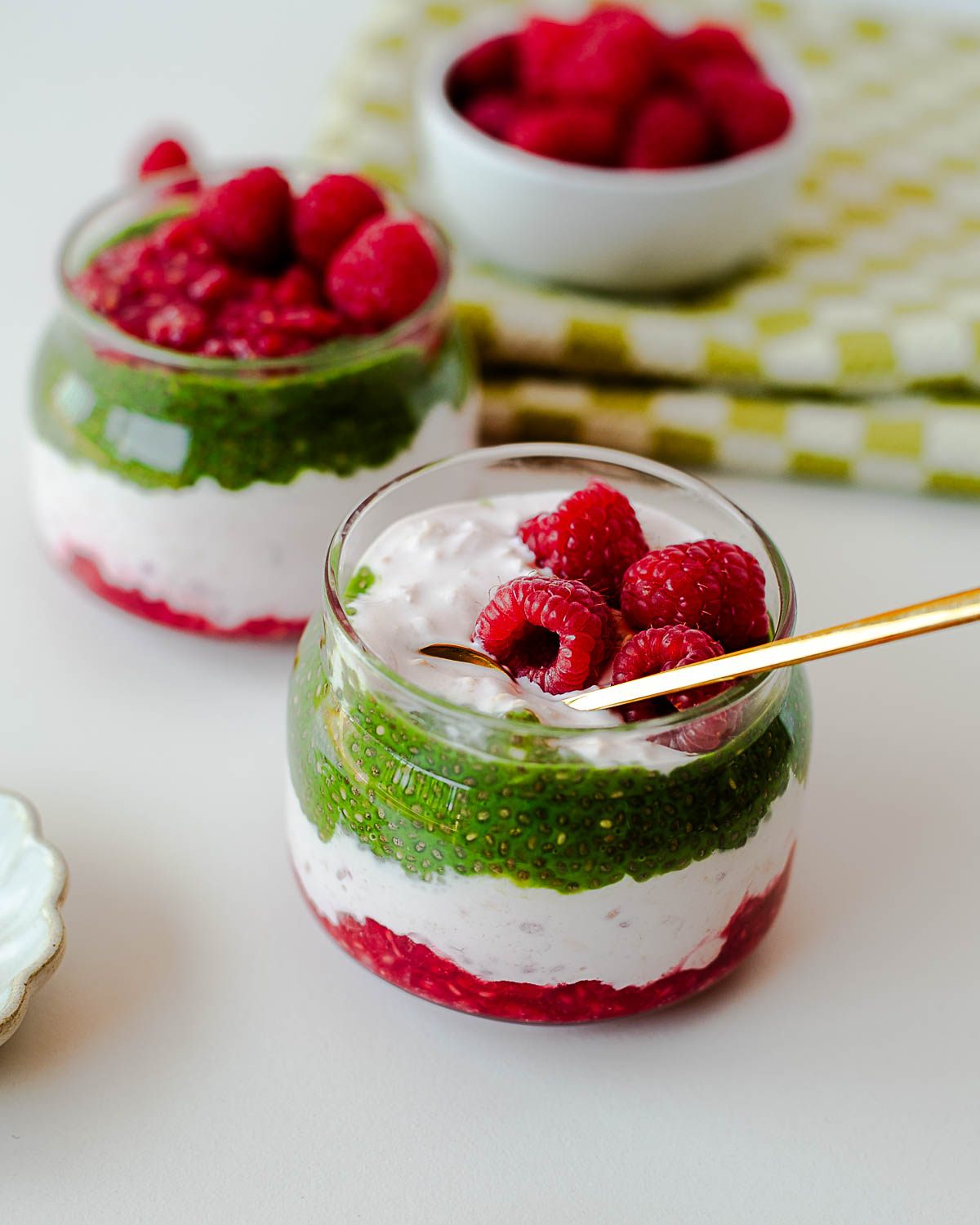 Styled raspberry matcha overnight oats parfaits arranged on a white background with fresh raspberries and kitchen linens.