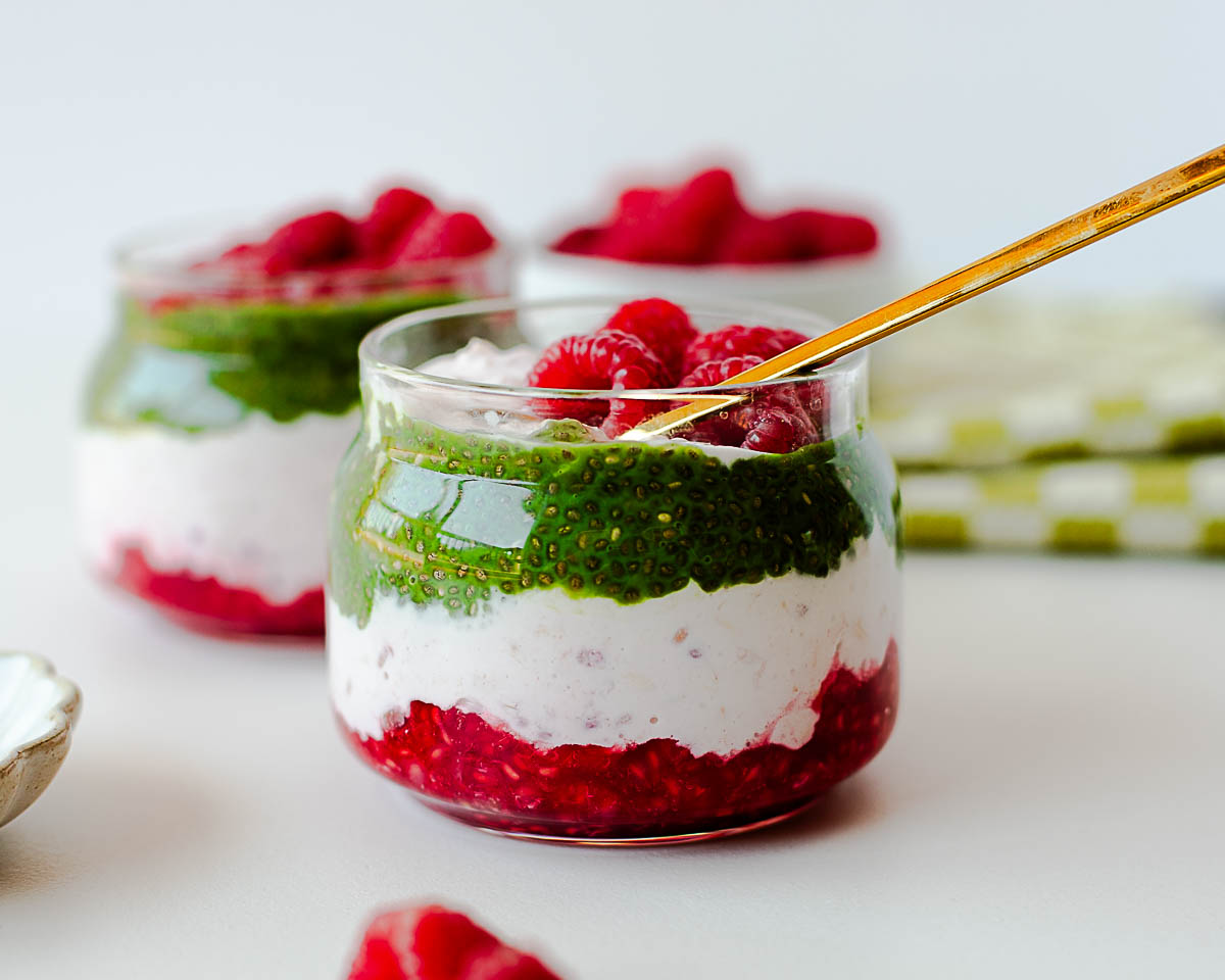Close-up of raspberry overnight oats parfait with matcha chia pudding layers topped with fresh raspberries and a gold spoon.