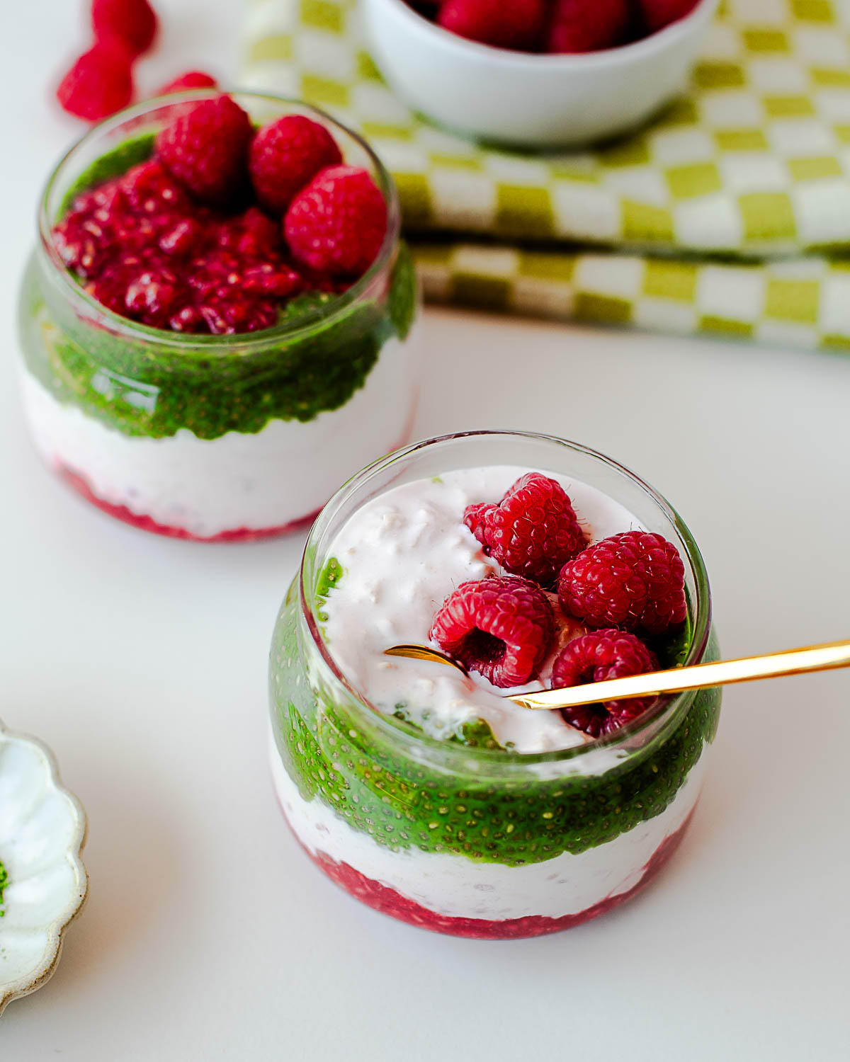 Two raspberry matcha overnight oats parfaits in glass jars with visible layers of chia pudding, yogurt, and raspberries.