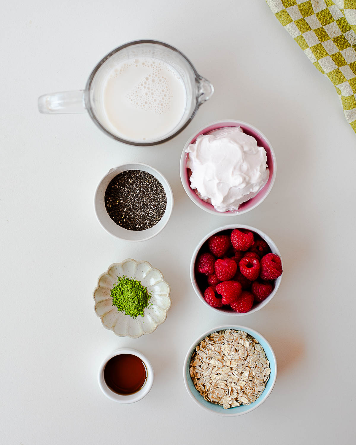 Ingredients for raspberry overnight oats and matcha chia pudding including rolled oats, raspberries, yogurt, chia seeds, matcha powder, maple syrup, and non-dairy milk arranged on a white surface.