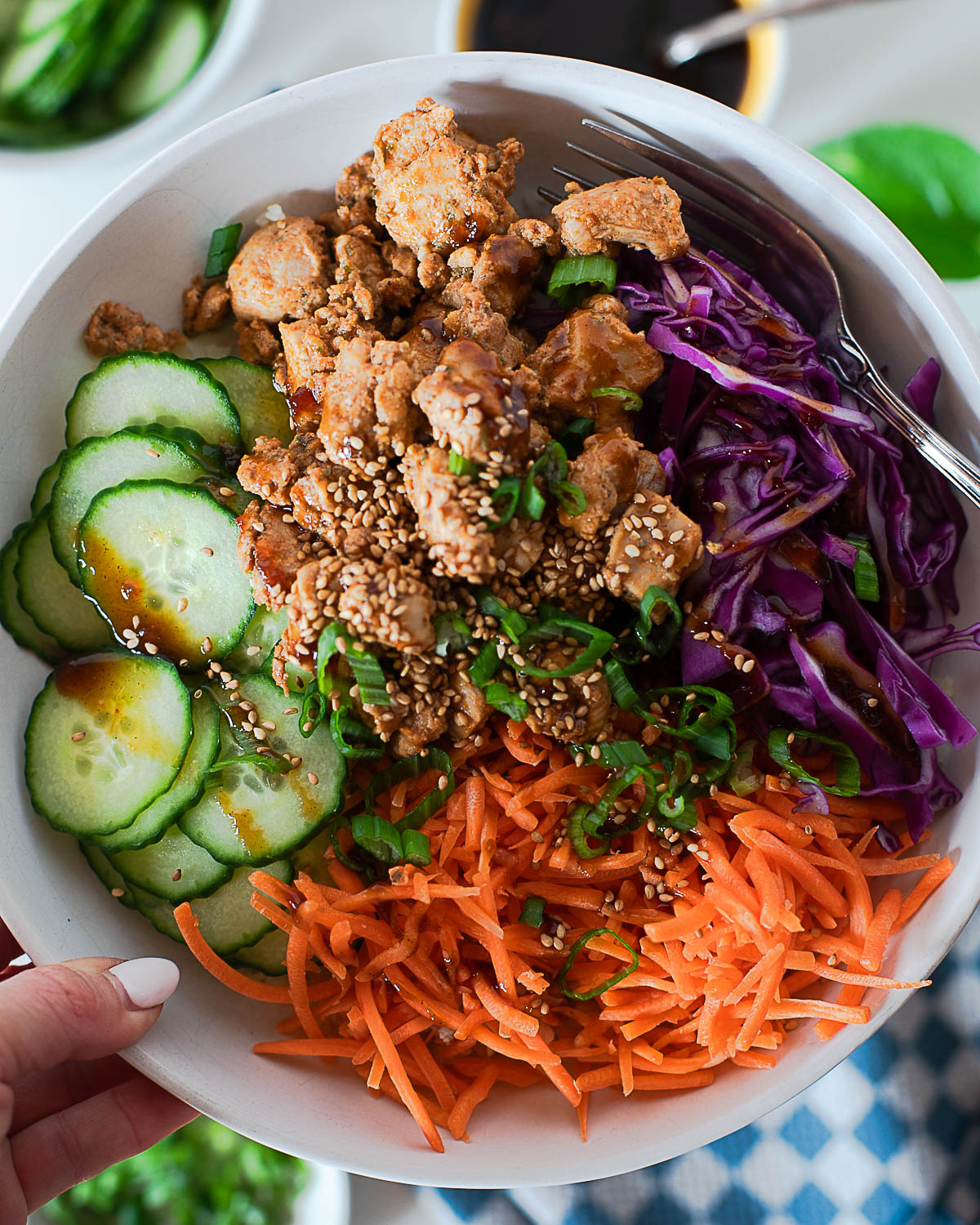 Hand holding a Korean BBQ chicken bowl with shredded carrots, cucumbers, cabbage, and saucy chicken.