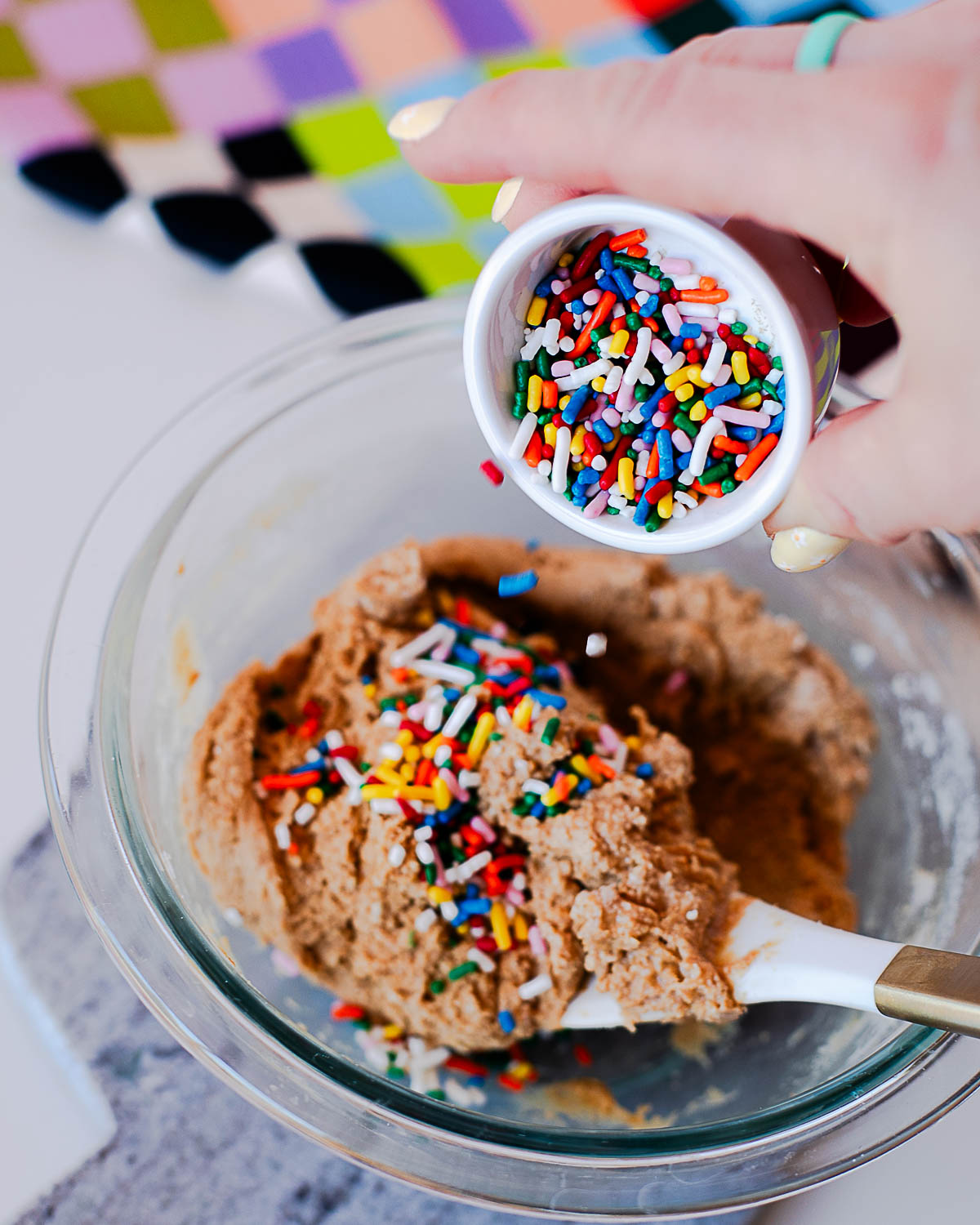 Hand pouring rainbow sprinkles into birthday cake protein ball dough in a mixing bowl.