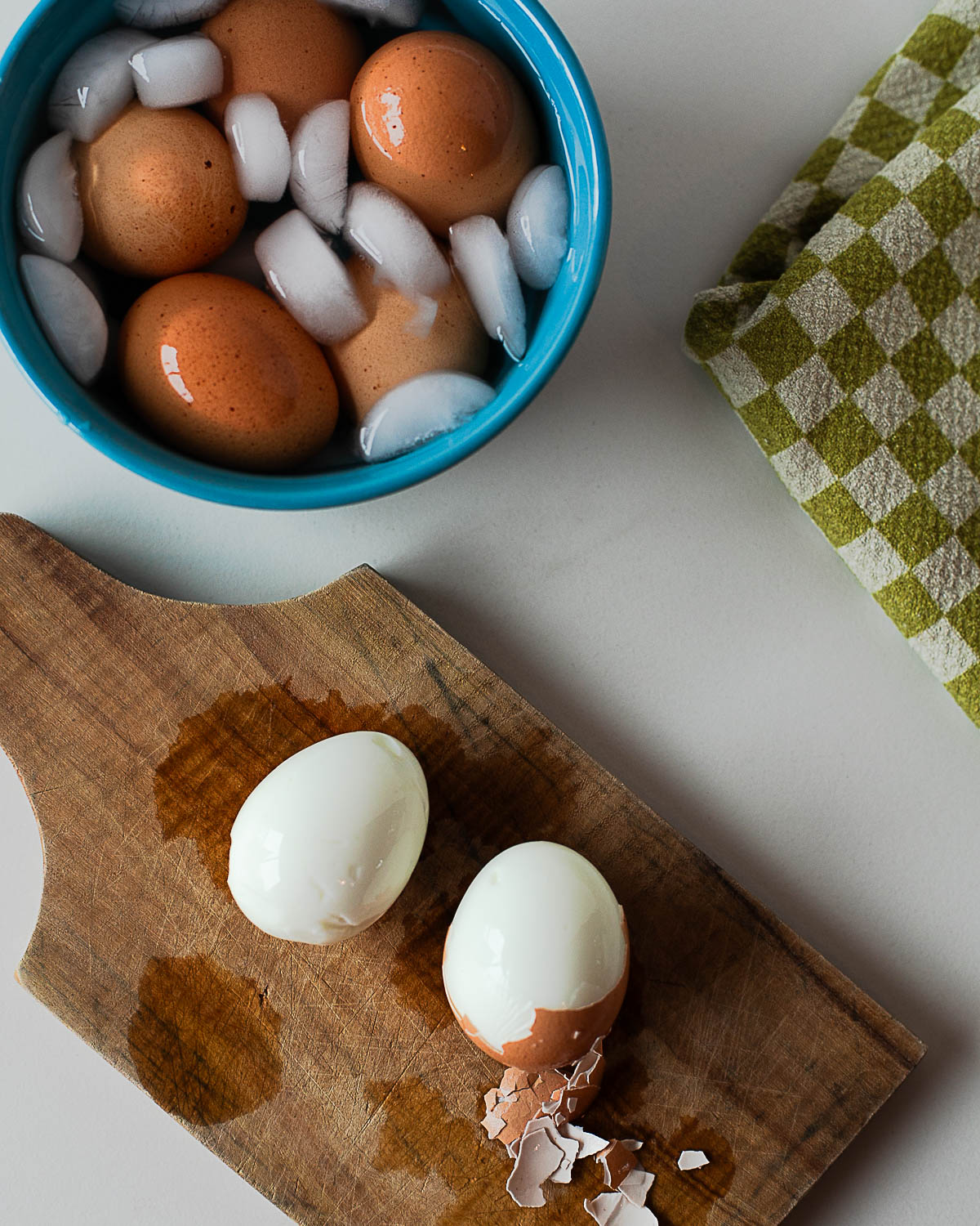 Hard-boiled eggs cooling in an ice bath in a blue bowl with ice cubes, next to a peeled egg on a wooden board