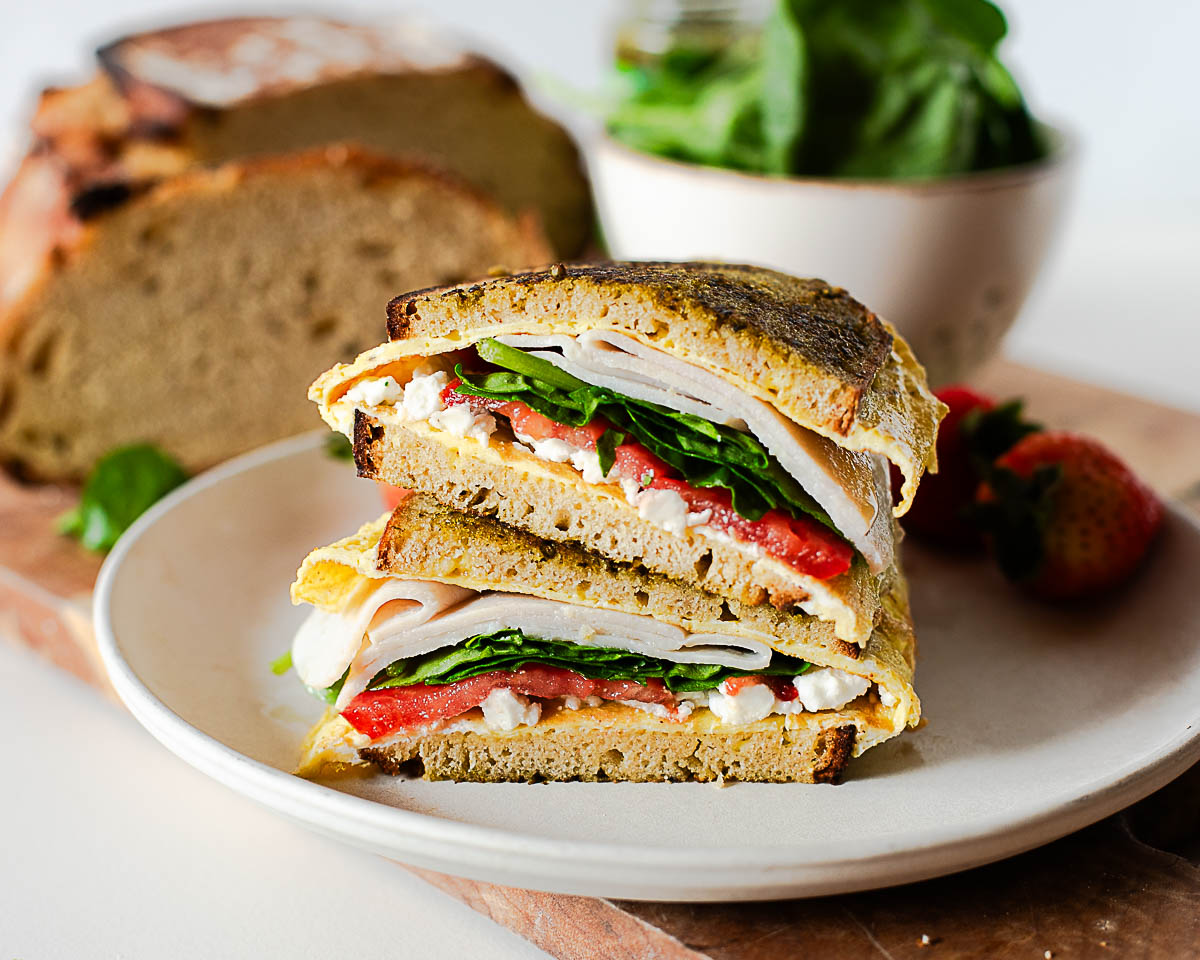 Close-up of pesto egg sandwich with melted feta, deli turkey, spinach, and tomato layered on crispy toasted bread