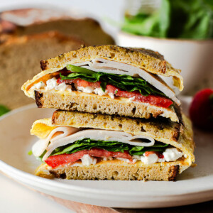 Pesto breakfast sandwich with eggs, feta, turkey, spinach, and tomato on toasted einkorn bread, sliced and stacked on a plate