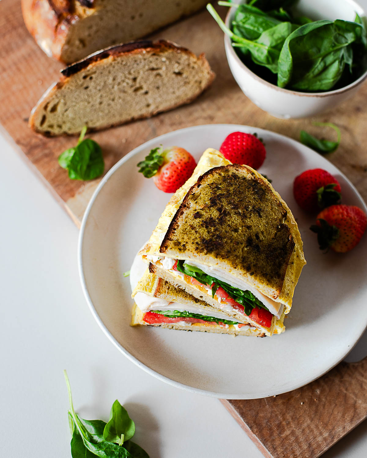 Top view of pesto breakfast sandwich on toasted bread with eggs, turkey, spinach, and tomato served on a plate with fresh strawberries