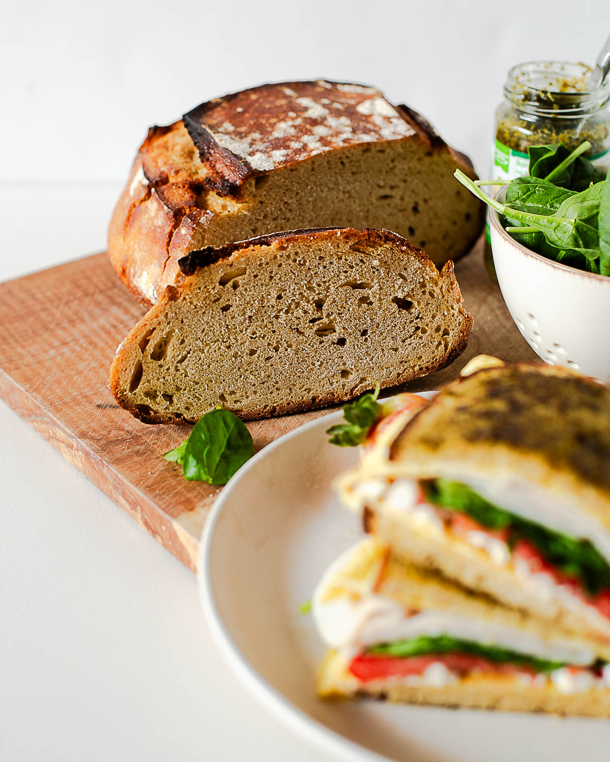 Sliced einkorn bread loaf with pesto breakfast sandwich in foreground filled with eggs, feta, turkey, spinach, and tomato