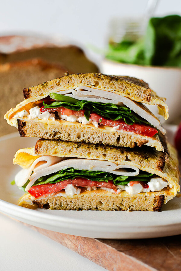 Pesto breakfast sandwich with eggs, feta, turkey, spinach, and tomato on toasted einkorn bread, sliced and stacked on a plate