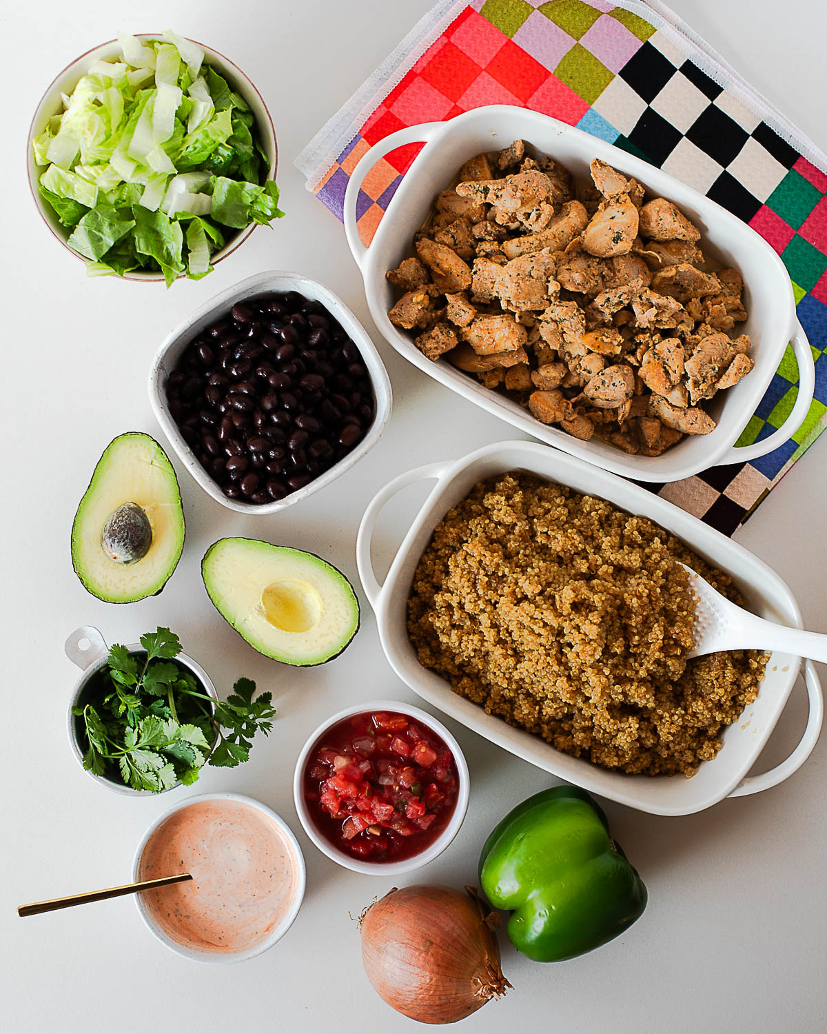 Overhead of southwest chicken burrito bowl ingredients including cooked chicken, quinoa, black beans, avocado, lettuce, pico de gallo, cilantro, and spicy ranch.