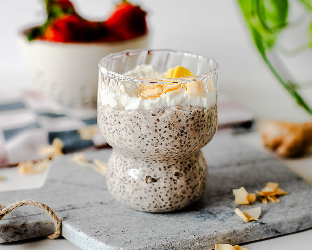 High protein coconut chia pudding in a glass cup topped with Greek yogurt and toasted coconut, styled with fresh strawberries and ginger in the background.