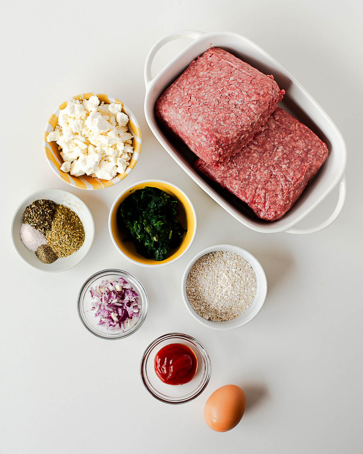 Ingredients for lamb burgers with feta including ground lamb, spinach, oat flour, herbs, egg, ketchup, and feta cheese arranged on a table