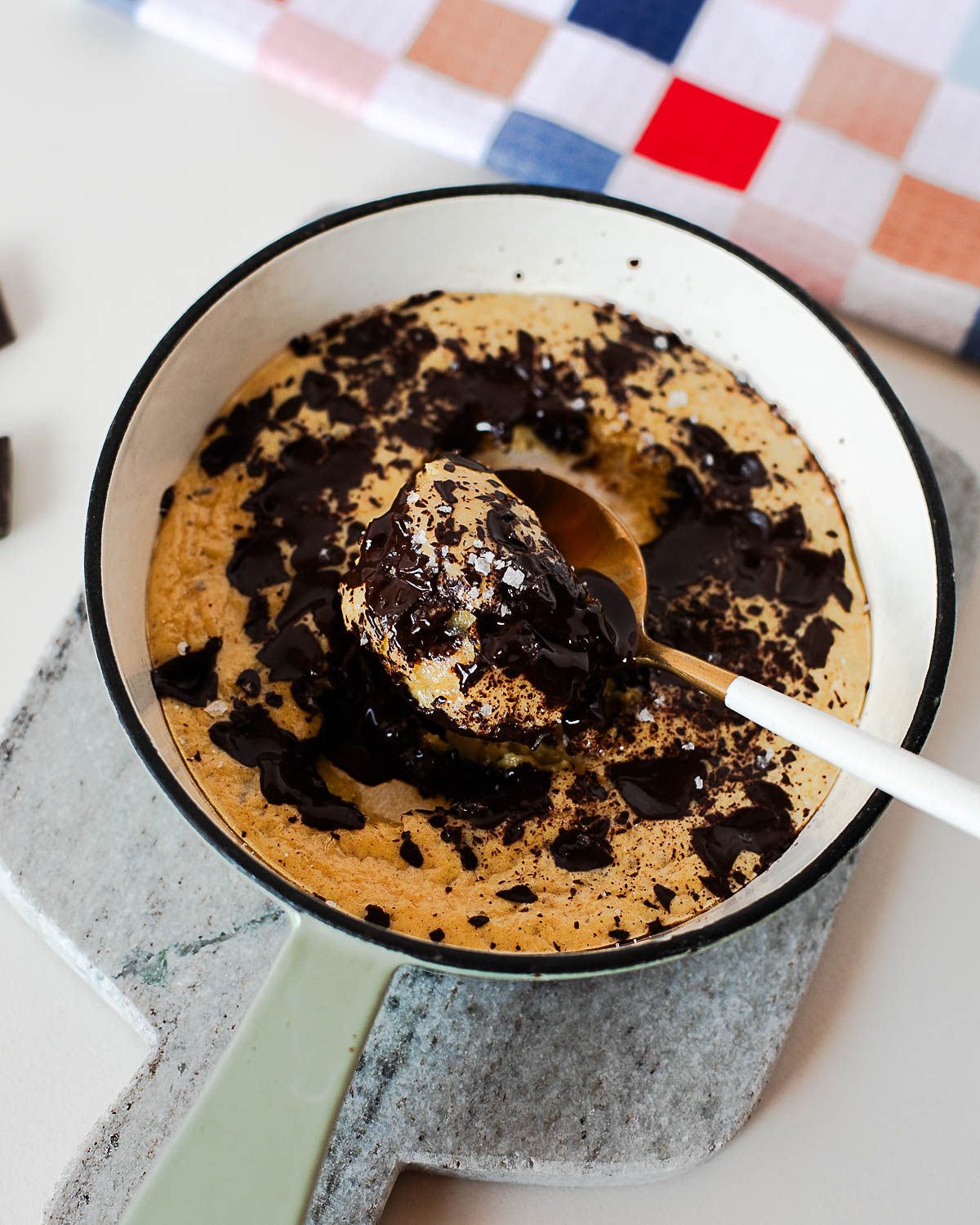 Spoon scooping into a gooey protein cookie skillet with melted dark chocolate and flaky sea salt on top.