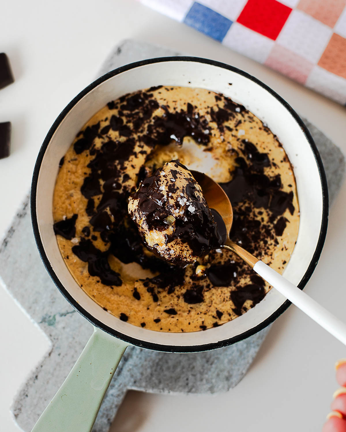 Spoon scooping into a gooey protein cookie skillet with melted dark chocolate and flaky sea salt on top.