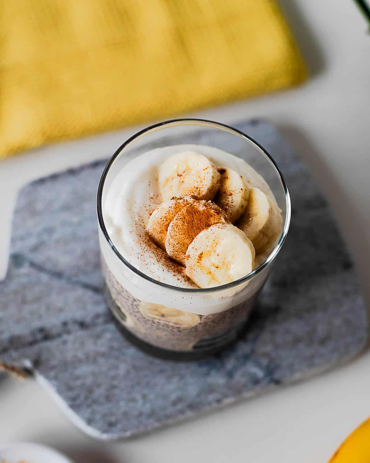 Overhead view of banana chia pudding topped with banana slices, creamy yogurt topping, and cinnamon in a clear serving glass.