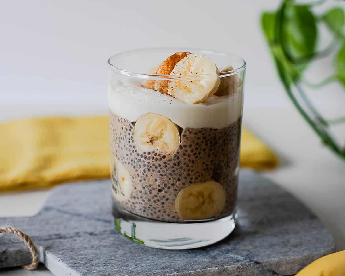 Banana chia pudding layered with a creamy protein yogurt topping and fresh banana slices in a clear glass, finished with a sprinkle of cinnamon.