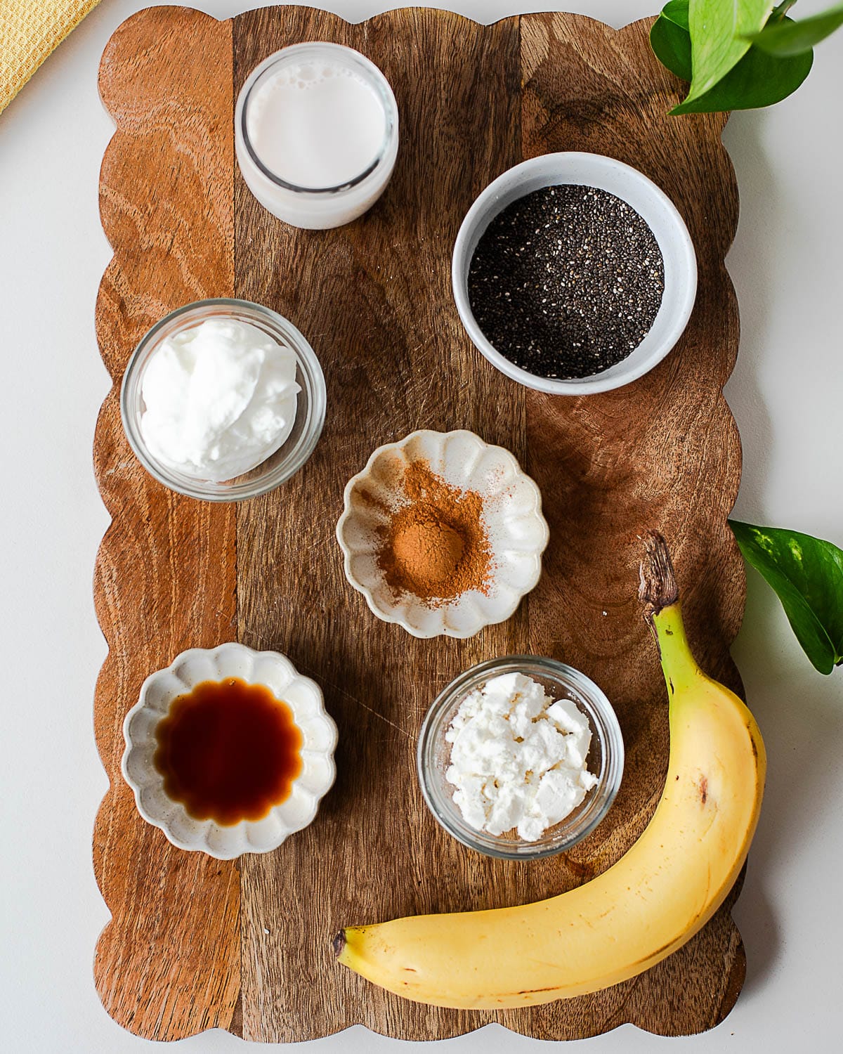 Ingredients for banana chia pudding including banana, chia seeds, Greek yogurt, almond milk, vanilla extract, cinnamon, coconut oil, and protein powder arranged on a wooden board.