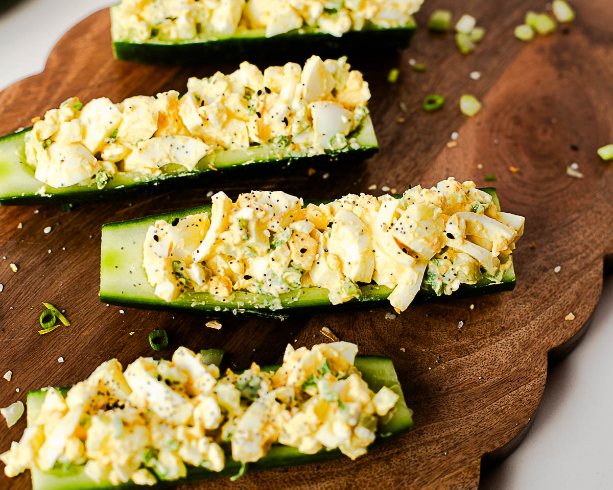Close-up of low-carb egg salad cucumber boats served on a wooden board with fresh cucumber and green onion garnish.