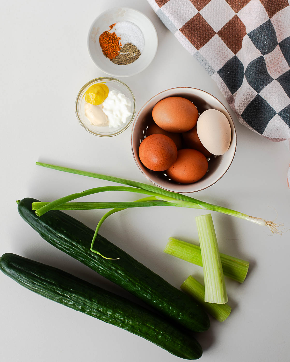Ingredients for egg salad cucumber boats including hard-boiled eggs, Greek yogurt, mayonnaise, mustard, celery, green onions, and English cucumbers arranged on a countertop.