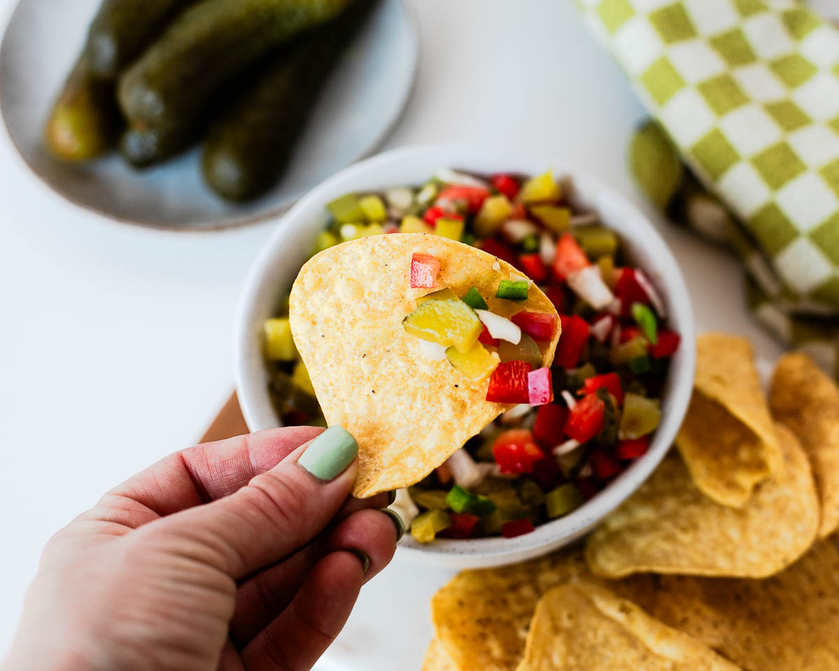 Tortilla chip dipped into homemade pickle de gallo, featuring diced dill pickles, bell pepper, onion, and jalapeño in every bite.