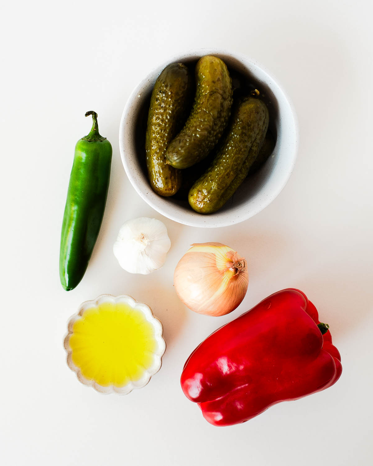 Ingredients for pickle de gallo arranged on a white surface, including dill pickles, red bell pepper, white onion, jalapeño, garlic, and pickle juice.