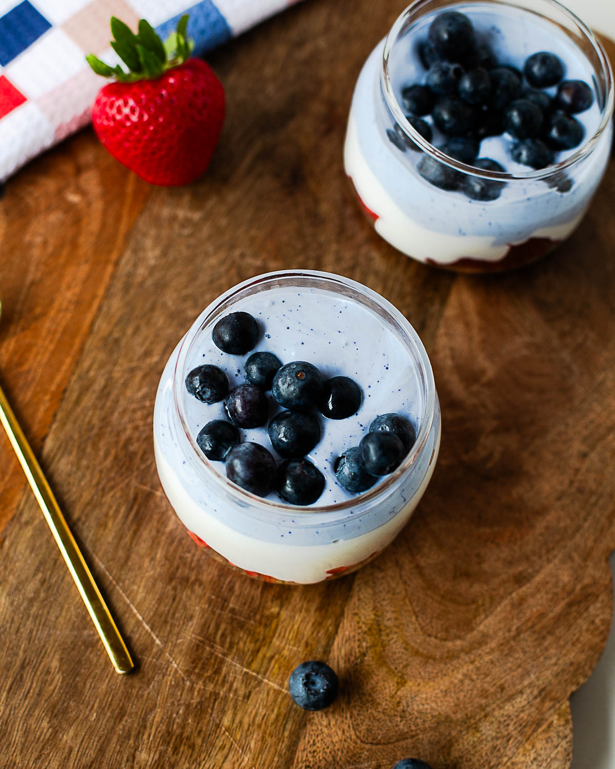 Overhead view of two patriotic cottage cheese parfaits topped with fresh blueberries on a wooden serving board with strawberries and a gold spoon.