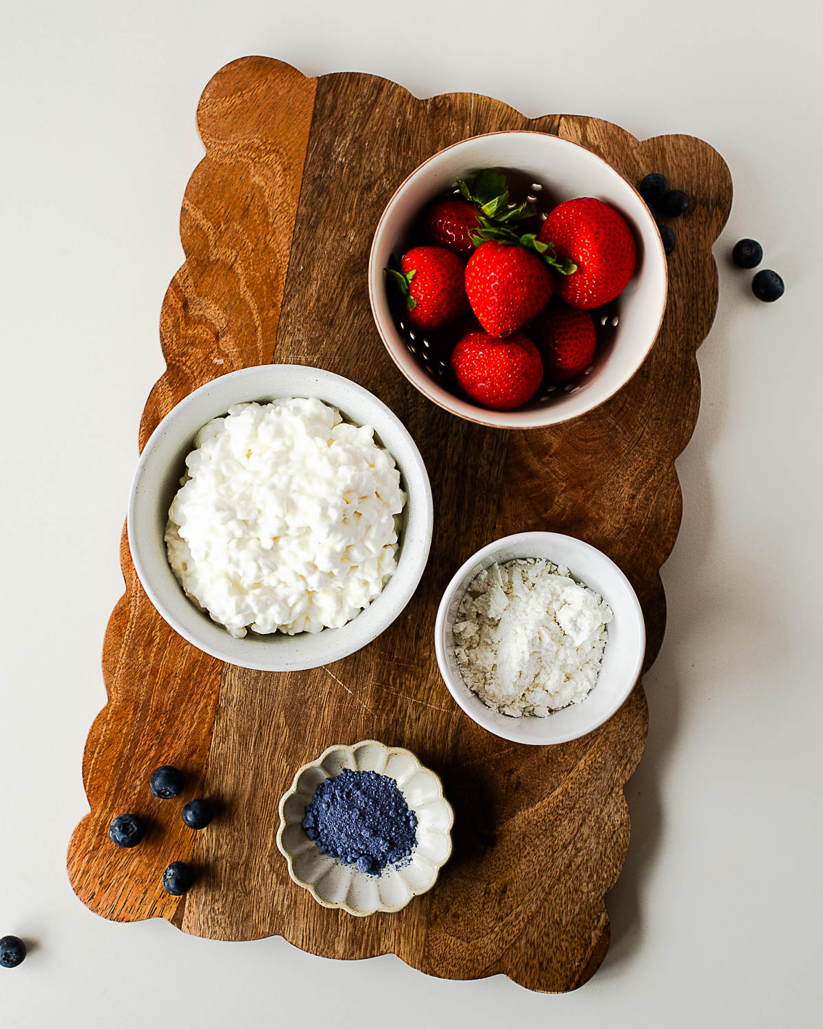 Ingredients for a high-protein cottage cheese parfait including cottage cheese, vanilla protein powder, butterfly pea flower powder, fresh strawberries, and blueberries on a wooden board.