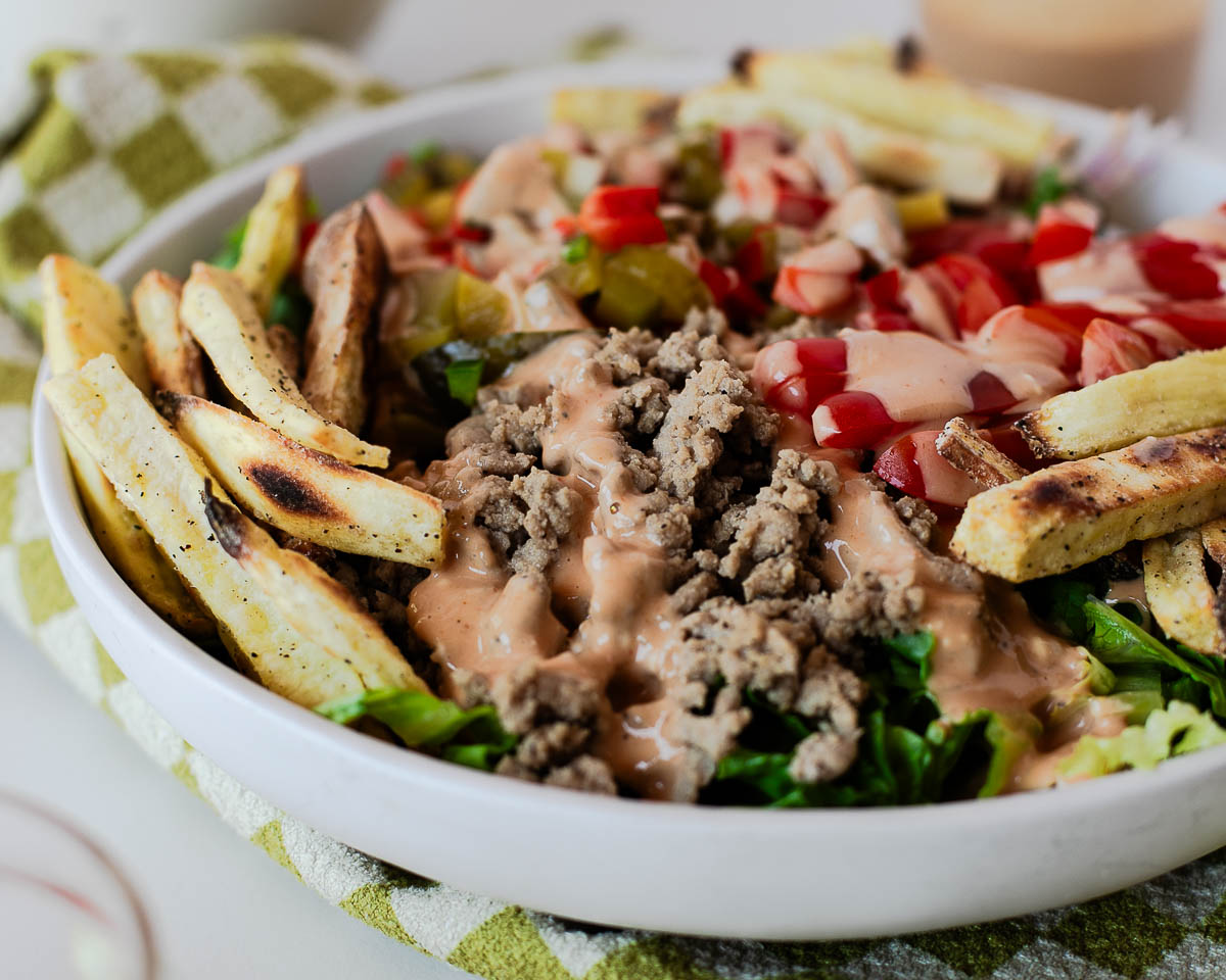 Close-up of a high-protein turkey burger bowl topped with homemade special sauce, fresh vegetables, pickle de gallo, and crispy sweet potato fries.