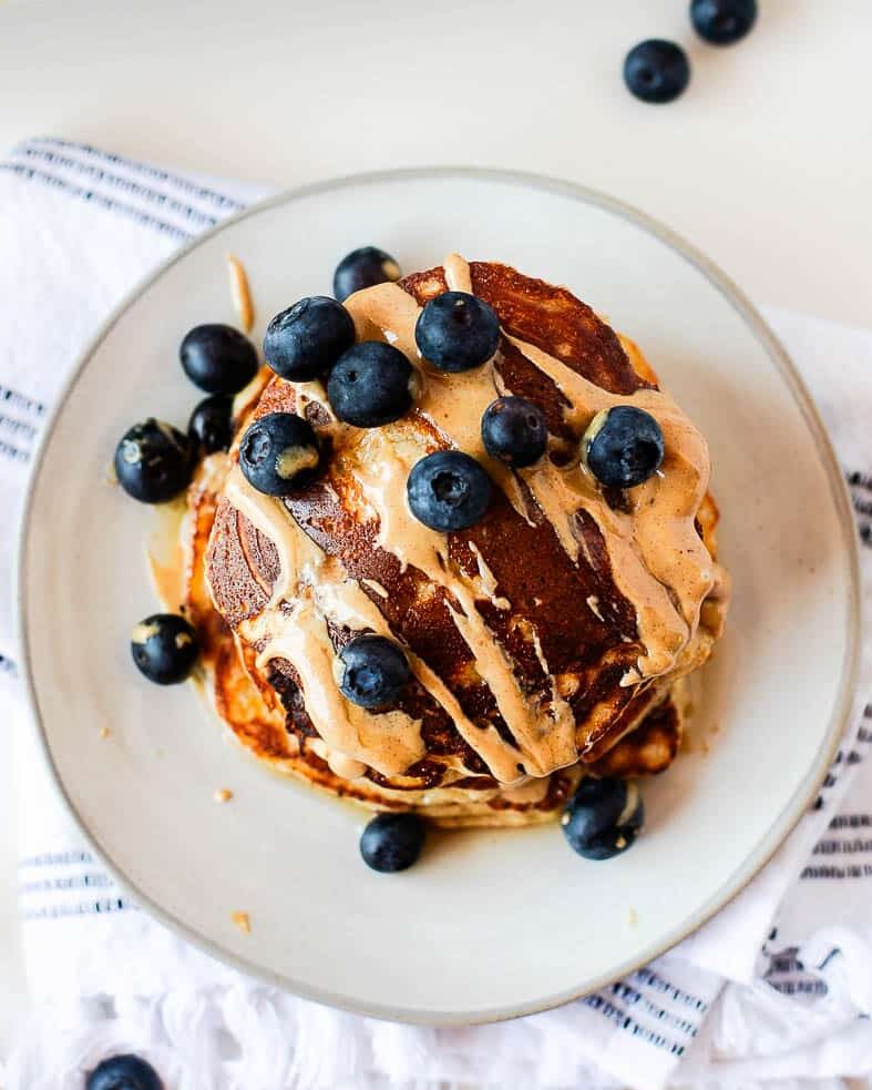Stack of fluffy banana coconut flour pancakes topped with fresh blueberries, peanut butter drizzle, and maple syrup on a white plate.
