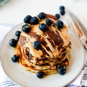 Close-up of fluffy coconut flour banana pancakes layered with fresh blueberries and drizzled with creamy peanut butter and maple syrup.