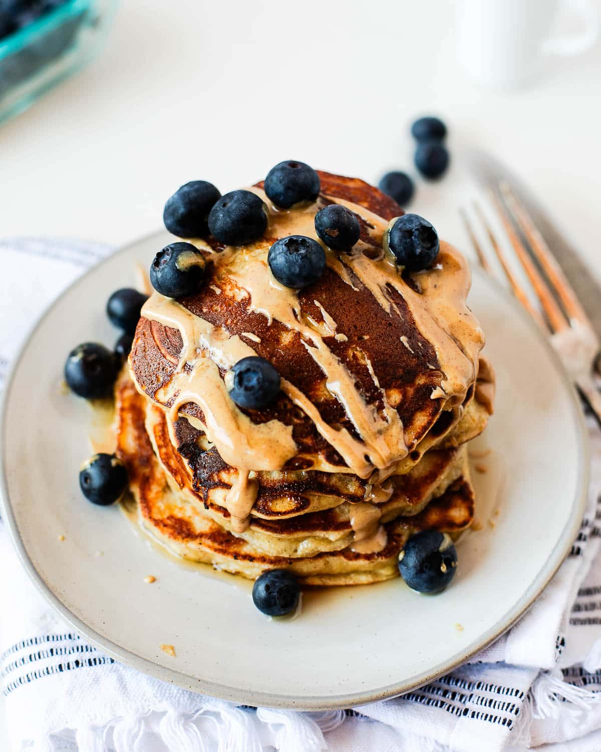 Close-up of fluffy coconut flour banana pancakes layered with fresh blueberries and drizzled with creamy peanut butter and maple syrup.