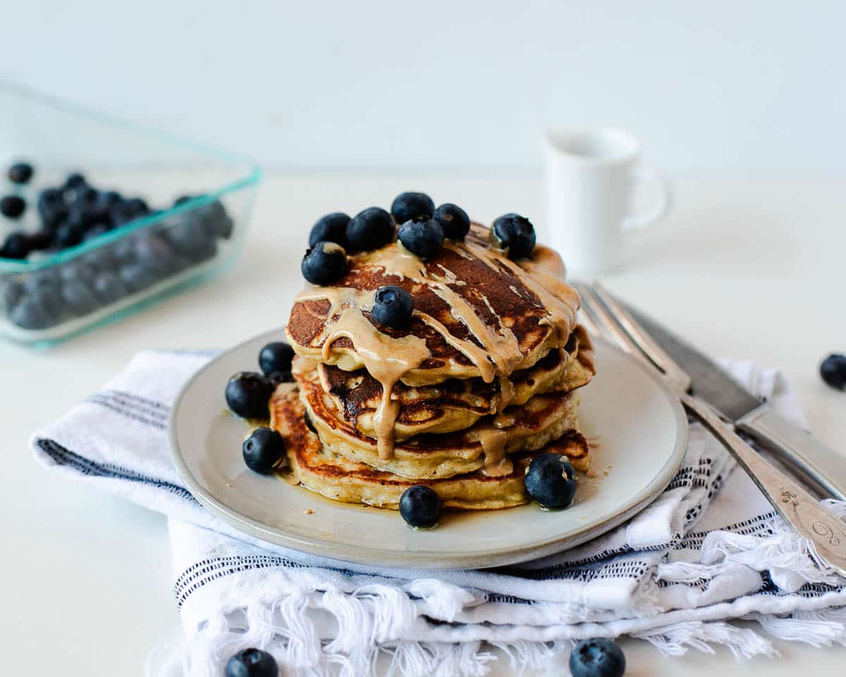 Blueberry banana coconut flour pancakes served with peanut butter drizzle and fresh blueberries on a white plate with a bowl of blueberries in the background.