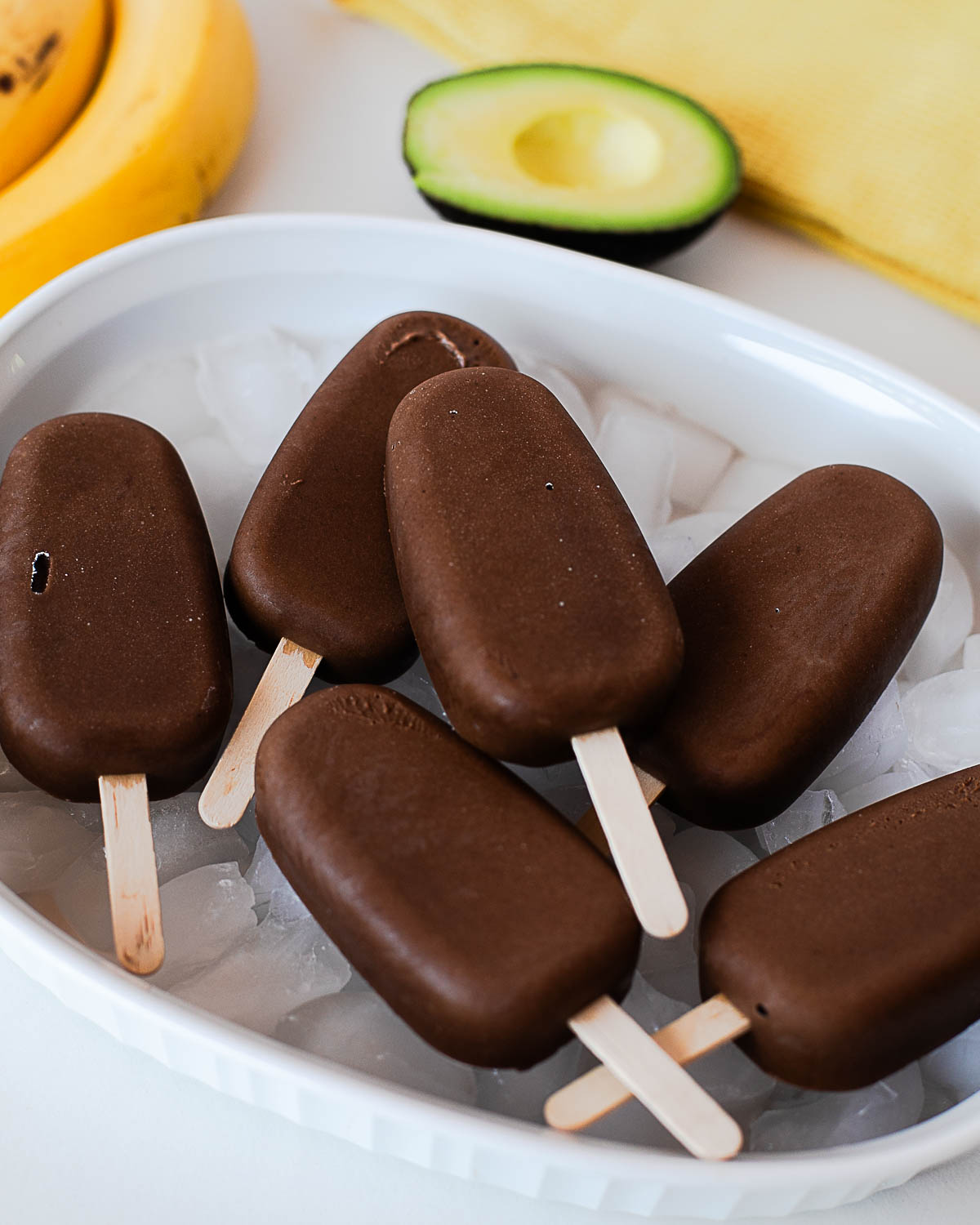 Six healthy chocolate avocado popsicles served over ice with fresh avocado and banana.